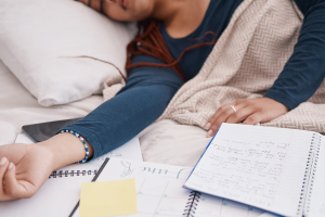 A woman lays sleeping on a bed surrounded by open notebooks and a calendar