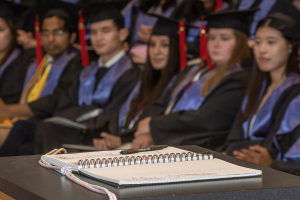 Graduates sit wearing caps and gowns in background with a open book on a tabletop in the foreground