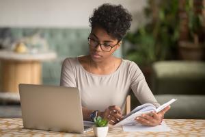 A women sits at a table holding a notebook and looking at a laptop screen
